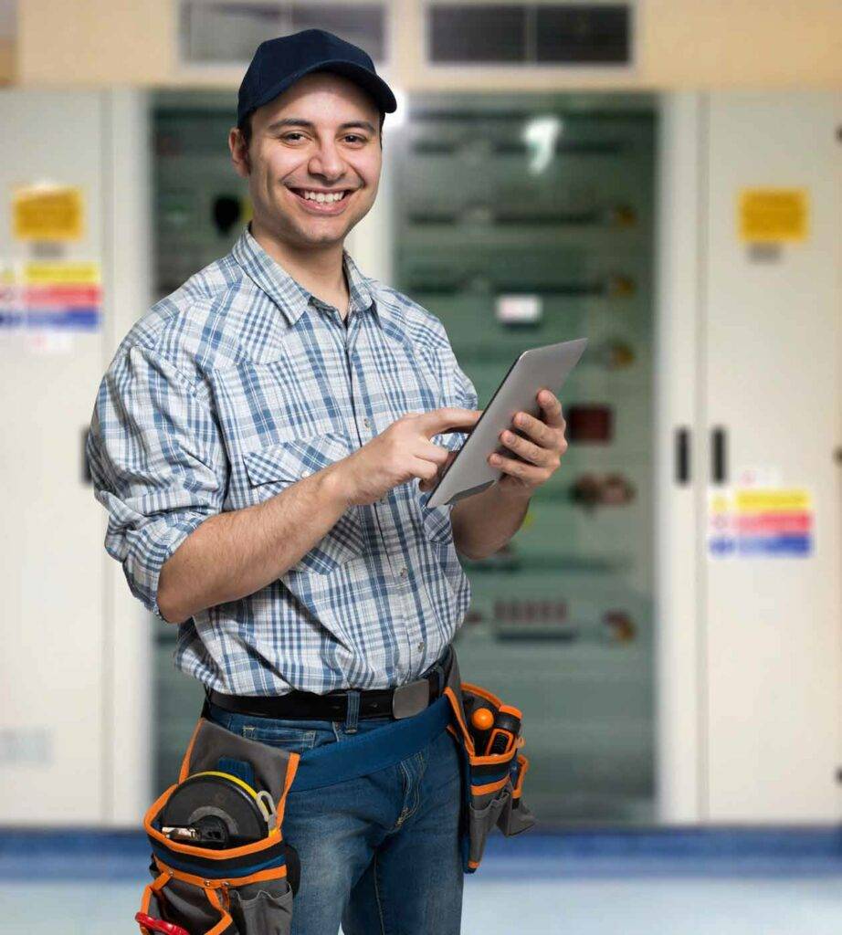 Freundlicher Hausmeister oder Elektriker mit Tablet steht vor einem Schaltschrank – Handwerkerjob an Schulen in Dresden