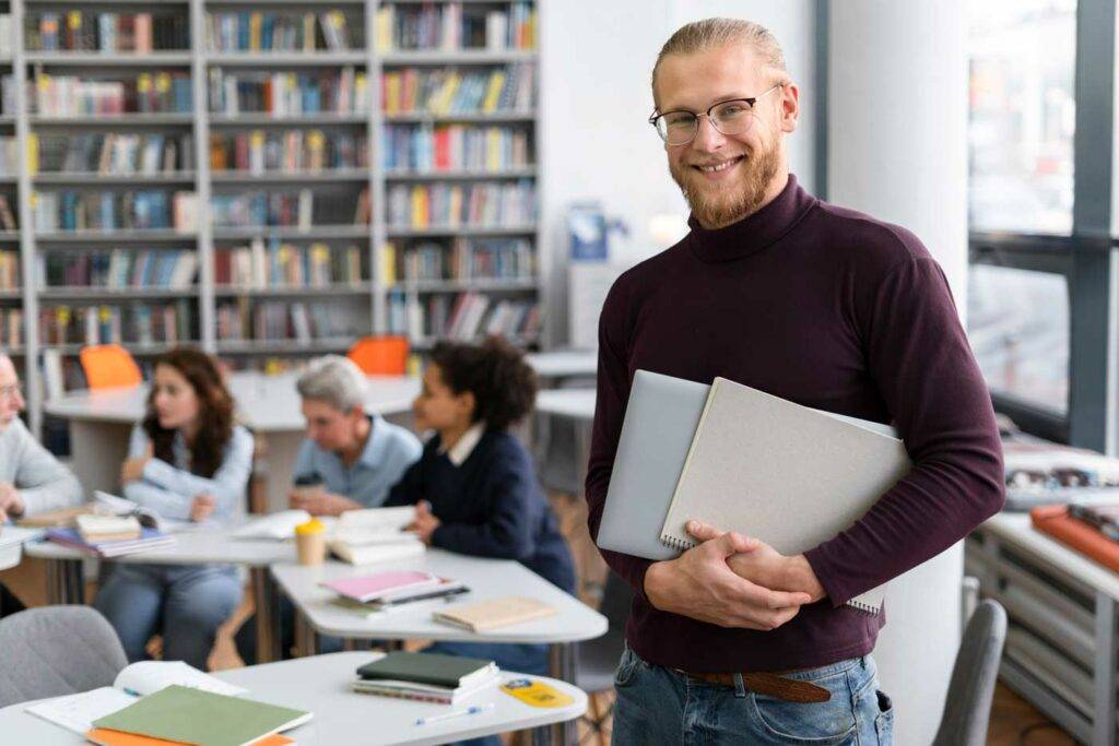 Erfahrener Dozent steht mit Ordnern in der Bibliothek eines Klassenzimmers und lächelt – Karriere und Jobs in Dresden